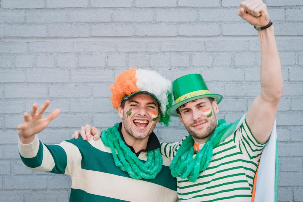 Joyful male friends in colorful traditional hats looking at camera while standing with raised arms near wall during Saint Patricks day